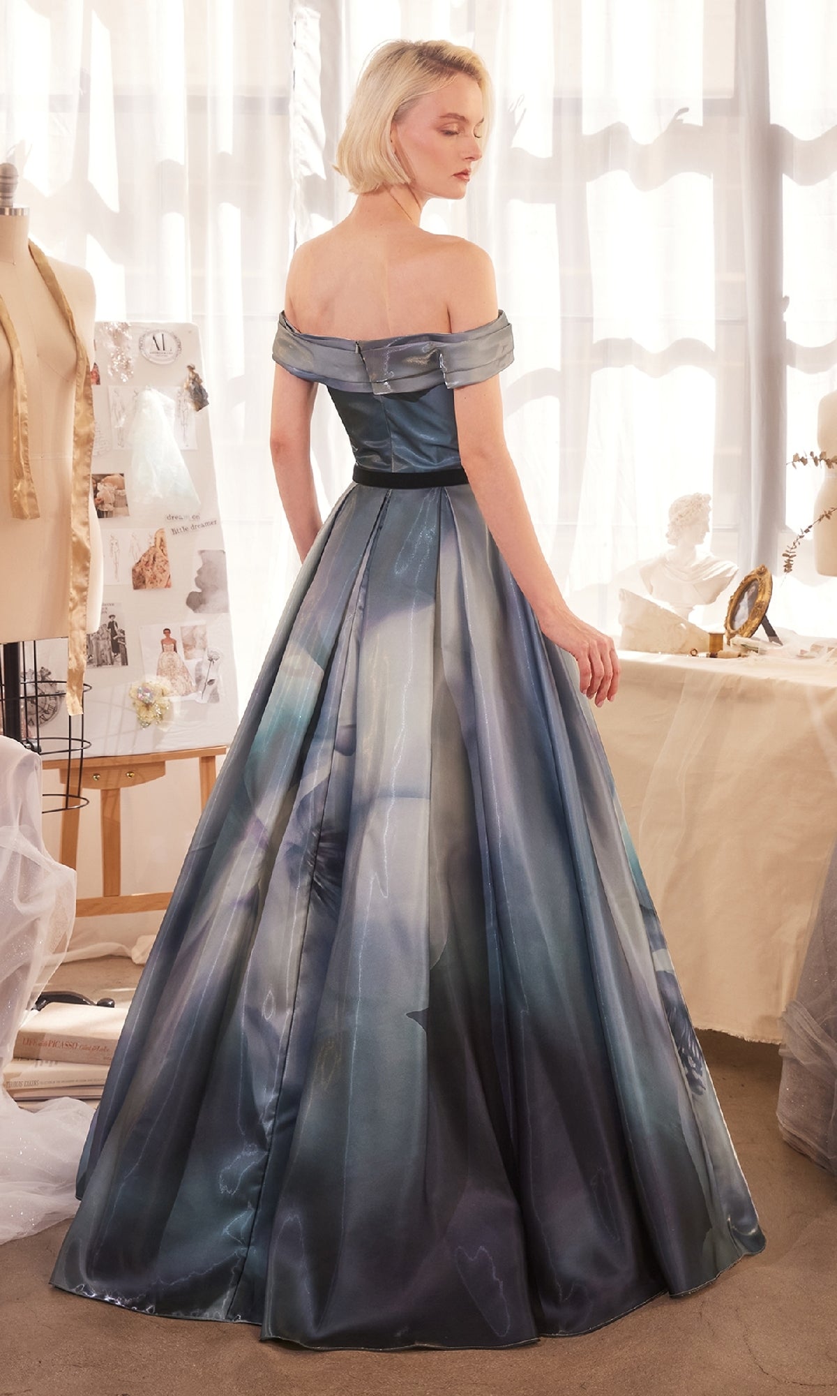 A woman with short blonde hair models the Off-Shoulder Blue Prom Gown: Andrea & Leo A1431, posing gracefully in a bright studio surrounded by sewing materials and dress forms.